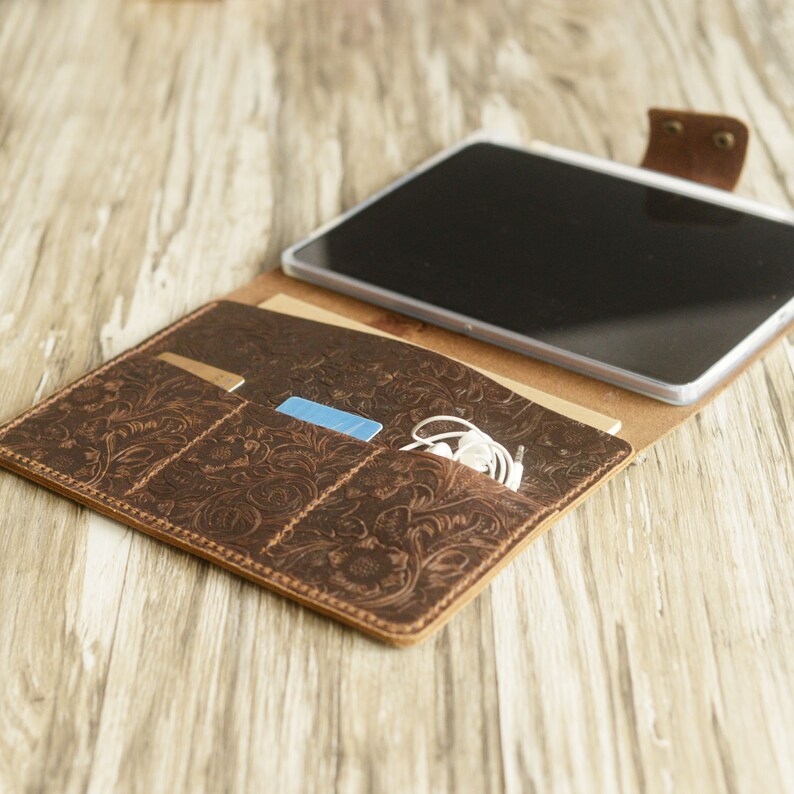 May include: A brown leather folio case with an embossed floral design. The case has three pockets for cards and a slot for a tablet or phone. White headphones are also visible in the case.