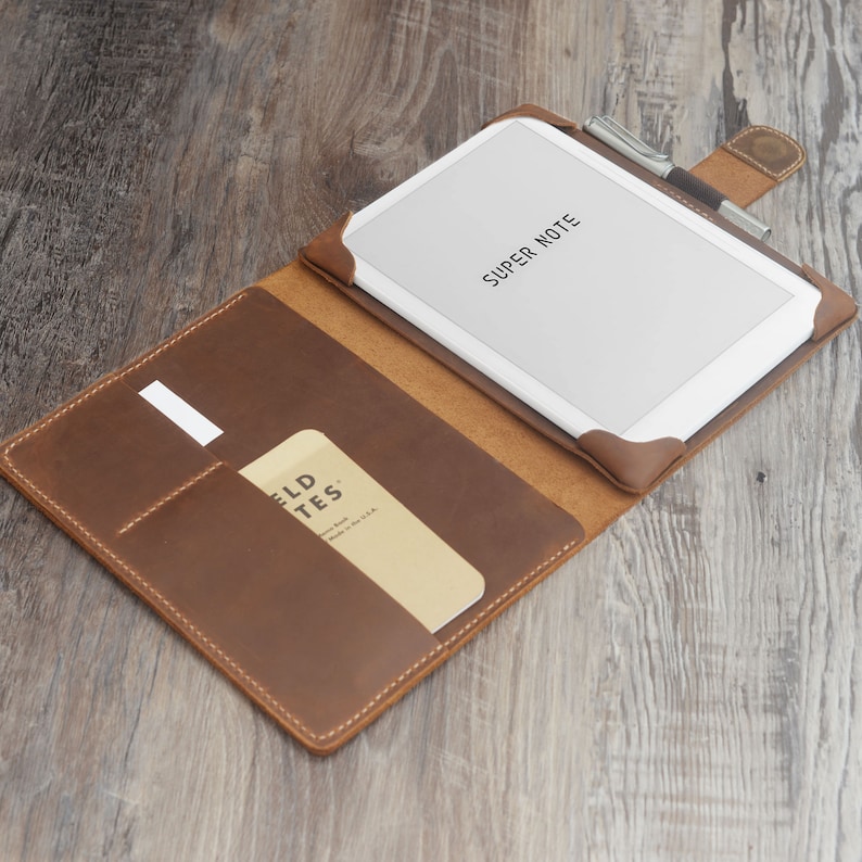 May include: A brown leather tablet case with a white tablet inside. The tablet displays the words "SUPER NOTE". A pen rests on the case, and a small notebook is in a pocket. The case is open, revealing the interior.