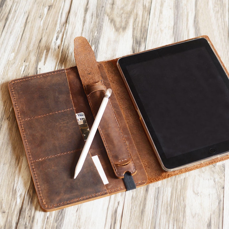 May include: A brown leather tablet case with a flap that folds over to protect the screen. The case has a slot for a stylus and a pocket for cards. A white stylus is inserted in the slot.