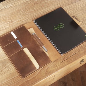 May include: A brown leather notebook cover with a black spiral bound notebook inside. The notebook has the text "Rocketbook" printed on the cover. The cover has multiple pockets and slots for holding cards, pens, and other items.