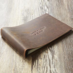 May include: A brown leather journal with the embossed text "Garrett & Lahna 8.23.2025". The journal is closed and has a curved design. The cover has a distressed appearance.