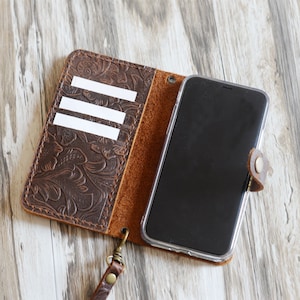 May include: A brown leather phone case with a floral embossed design. The case has a flap that folds over the phone and a slot for cards. The case also has a wrist strap.
