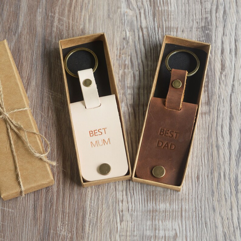 May include: Two leather keychains in boxes. One keychain is light brown with the text "BEST MUM" and the other is dark brown with the text "BEST DAD".