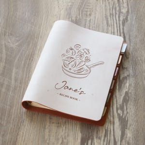 May include: A light-colored recipe book binder with a dark brown leather-like spine and edges. The front cover features a brown line-art illustration of vegetables in a frying pan, with the text Jane's RECIPE BOOK printed below the graphic.