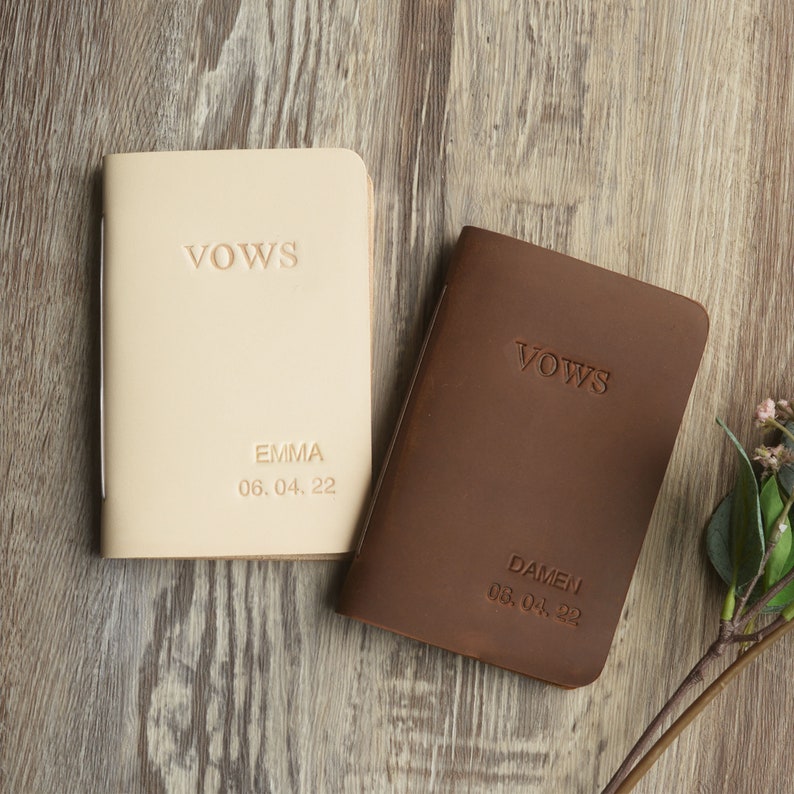 May include: Two leather-bound vow books, one beige and one brown, with the word "Vows" embossed on the front. The beige book has the name "Emma" and the date "06.04.22" embossed on the front. The brown book has the name "Damen" and the date "08.04.22" embossed on the front.