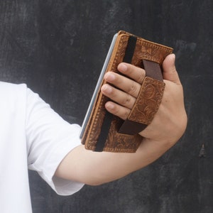 Peut inclure: Une coque de t&eacute;l&eacute;phone en cuir marron avec un motif floral et une sangle &eacute;lastique noire. La coque est dot&eacute;e d'une dragonne pour un transport facile.