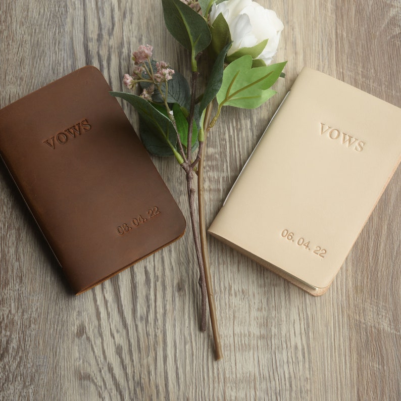 May include: Two leather-bound vow books, one brown and one beige, with the word "Vows" embossed on the front. The date is embossed on the bottom of each book: 08.04.22 and 06.04.22.