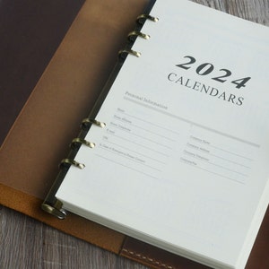 May include: A white, leather-bound planner with a gold ring binding. The cover has the text "2024 Calendars" and "Personal Information" with blank lines for filling in personal details.