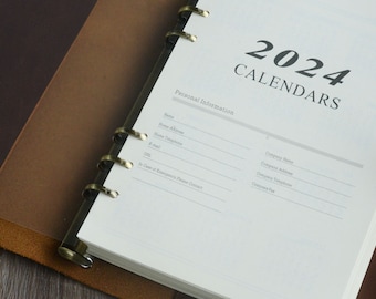 May include: A white, leather-bound planner with a gold ring binding. The cover has the text "2024 Calendars" and "Personal Information" with blank lines for filling in personal details.