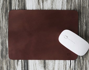 May include: A brown leather mouse pad with a white wireless mouse on top of it. The mouse pad has a small logo in the bottom right corner.