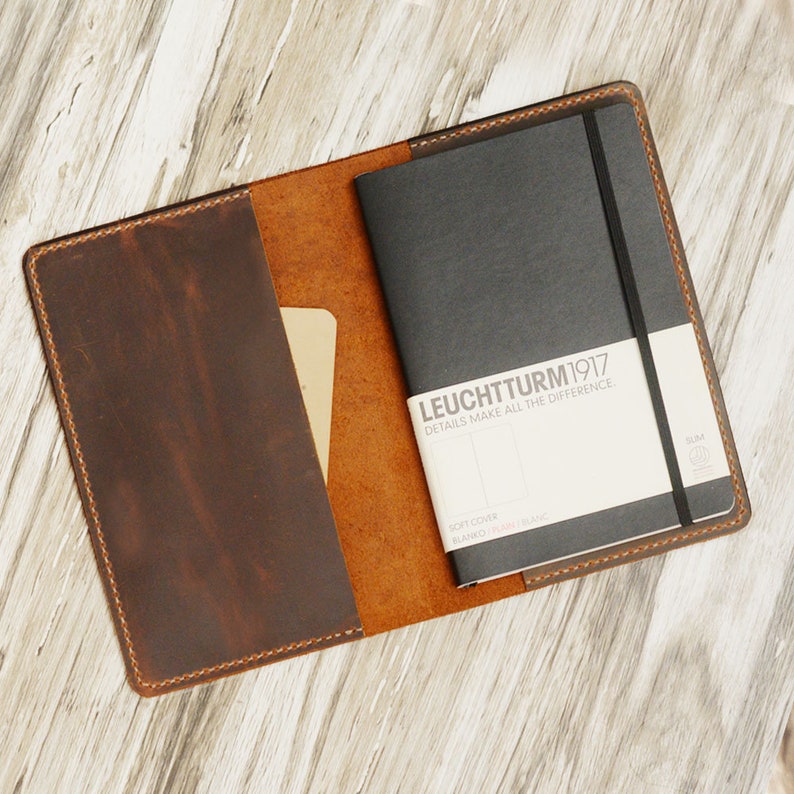 May include: A brown leather notebook cover with a black notebook inside. The notebook has a black elastic band and a white label that says "LEUCHTTURM1917 DETAILS MAKE ALL THE DIFFERENCE."