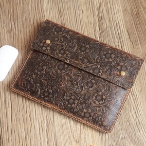Peut inclure: Une pochette pour ordinateur portable en cuir marron avec un motif en relief floral. La pochette a une fermeture &agrave; rabat avec un bouton en laiton.