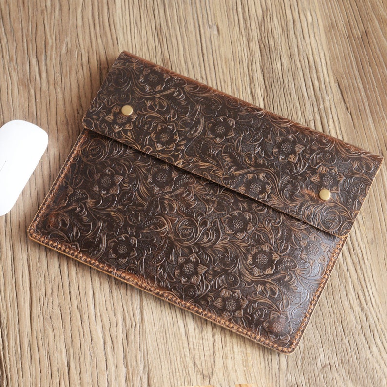 Peut inclure: Une pochette pour ordinateur portable en cuir marron avec un motif en relief floral. La pochette a une fermeture &agrave; rabat avec un bouton en laiton.