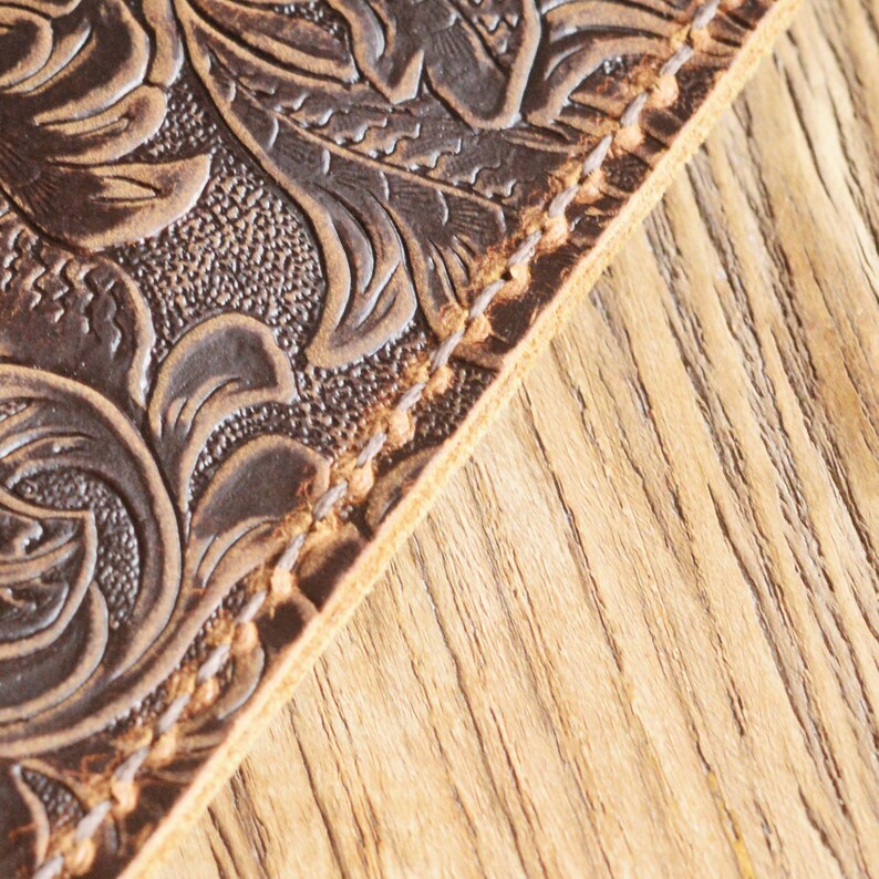 Peut inclure: Gros plan sur une surface en cuir marron avec un motif en relief floral. Le cuir est cousu le long du bord avec du fil marron.