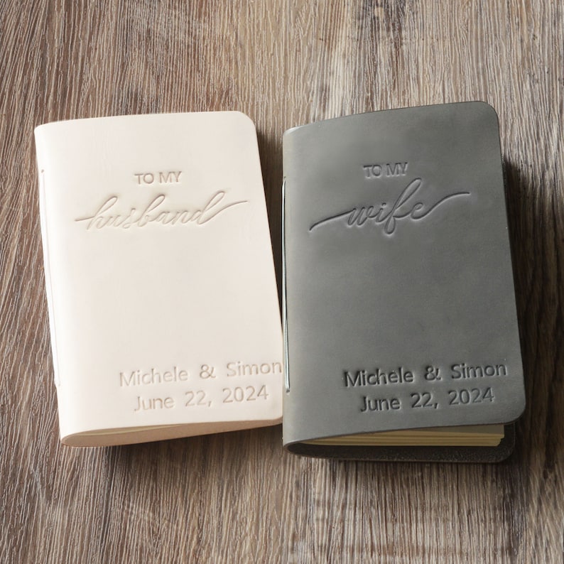 May include: Two leather-bound journals, one in beige and one in gray, with the words "To My Husband" and "To My Wife" embossed on the covers. Both journals have the names "Michele & Simon" and the date "June 22, 2024" printed on the bottom.