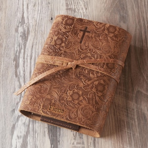 Może przedstawiać: Brązowy skórzany notatnik z wytłoczonym kwiatowym wzorem i symbolem krzyża. Notatnik jest związany skórzanym paskiem i ma imię "Fiona" na okładce. Notatnik jest wykonany ze skóry.