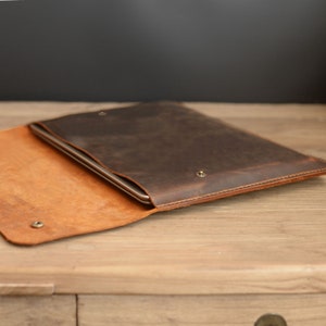 Puede incluir: Una funda para port&aacute;til de cuero marr&oacute;n con una solapa que se abre para revelar un interior de cuero beige. La funda est&aacute; hecha de cuero envejecido y tiene un brillo sutil.