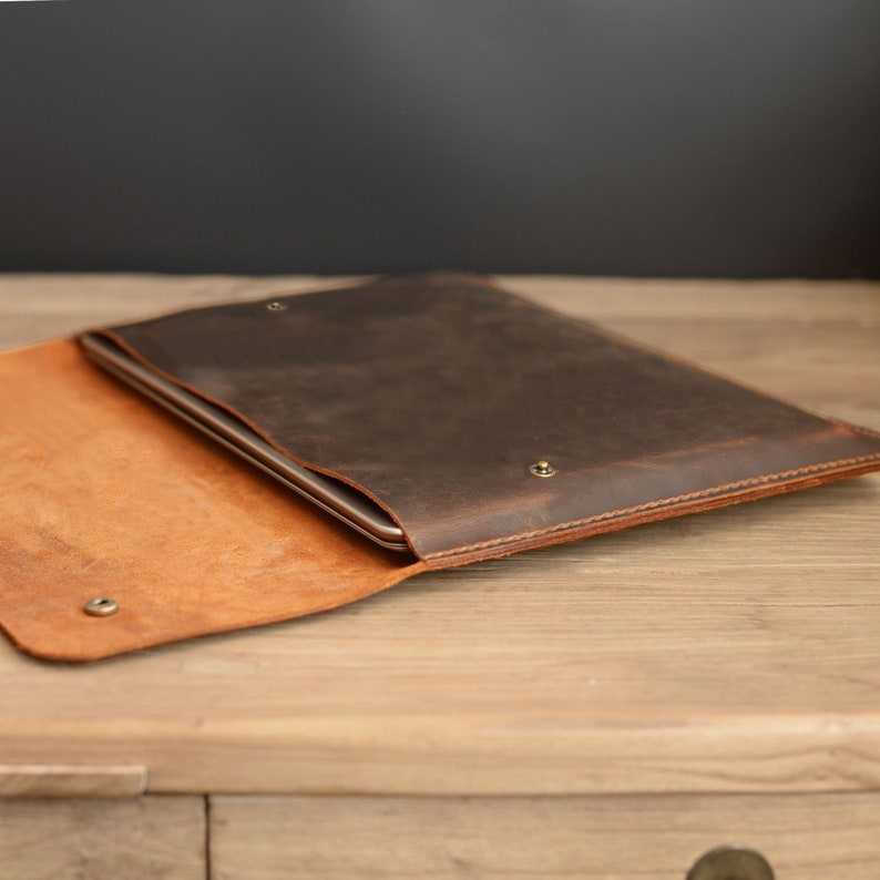 Puede incluir: Una funda para port&aacute;til de cuero marr&oacute;n con una solapa que se abre para revelar un interior de cuero beige. La funda est&aacute; hecha de cuero envejecido y tiene un brillo sutil.