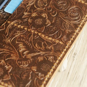May include: Close-up of a brown leather wallet with a floral embossed design. The wallet has a stitched edge and a subtle distressed look.