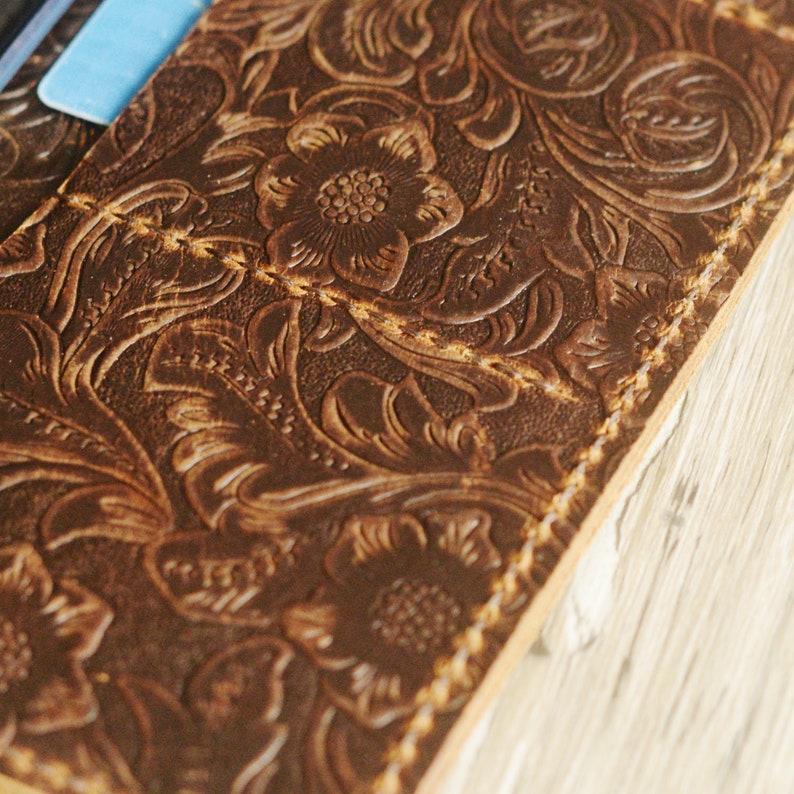 May include: Close-up of a brown leather wallet with a floral embossed design. The wallet has a stitched edge and a subtle distressed look.