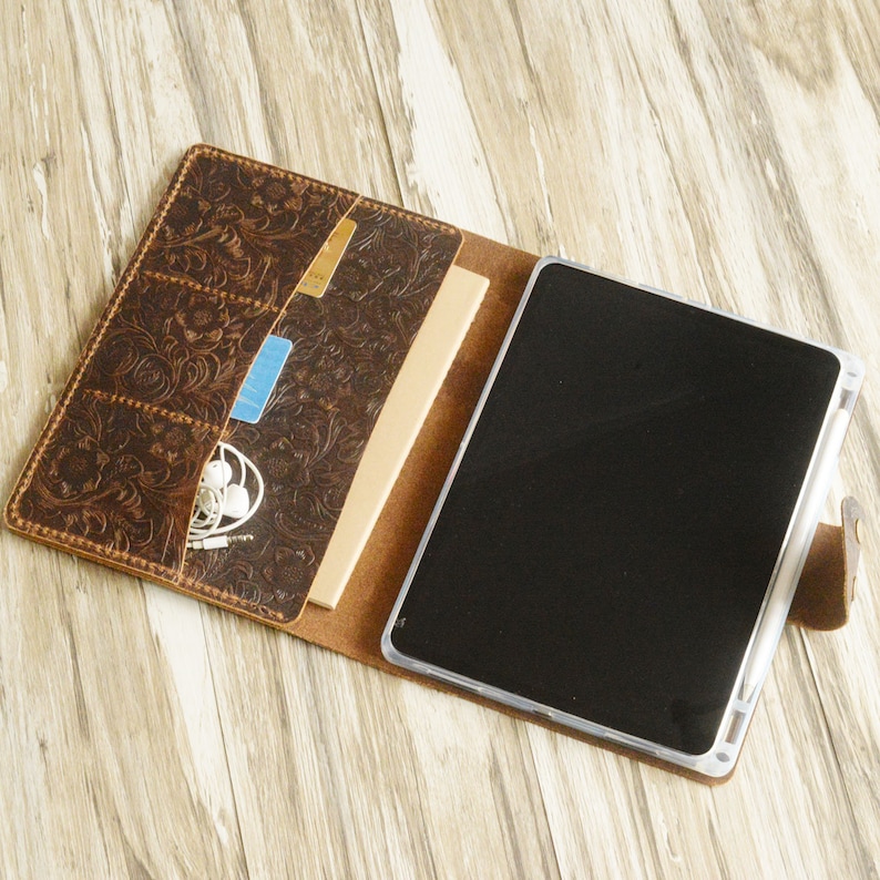May include: A brown leather folio case with an embossed floral design. The case is open, revealing a tablet and a notebook. There are pockets on the inside of the case for holding cards and other small items.