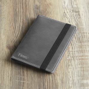Peut inclure: Un porte-passeport en cuir gris avec une fermeture &agrave; bande &eacute;lastique noire. Le porte-passeport porte le nom "Fiona" en relief sur le devant.