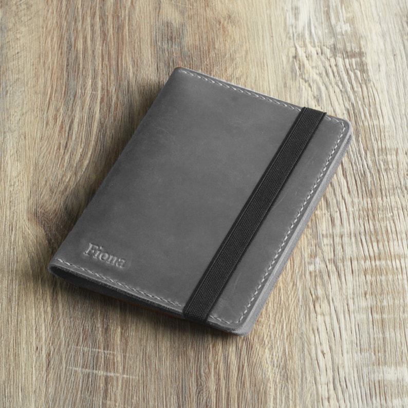 Peut inclure: Un porte-passeport en cuir gris avec une fermeture &agrave; bande &eacute;lastique noire. Le porte-passeport porte le nom "Fiona" en relief sur le devant.