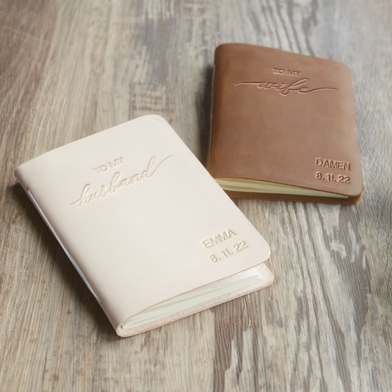 May include: Two leather-bound journals with personalized inscriptions. One journal is brown and says "To my wife" with the name "Damen" and the date "8.11.22". The other journal is cream colored and says "To my husband" with the name "Emma" and the date "8.11.22".