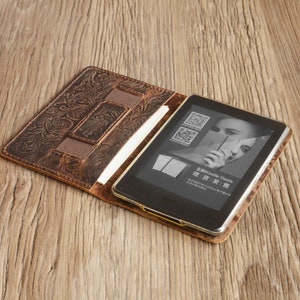Może przedstawiać: Brązowe sk&oacute;rzane etui na Kindle Oasis z tłoczonym wzorem kwiatowym. Etui posiada wbudowaną kieszeń na kartę kredytową lub inne małe przedmioty. Kindle Oasis jest widoczny wewnątrz etui, wyświetlając promocyjny obraz urządzenia.