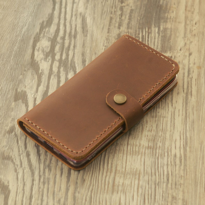 Peut inclure: Une coque de t&eacute;l&eacute;phone en cuir marron avec une fermeture &agrave; rabat et un bouton pression. La coque est en cuir v&eacute;ritable et a un aspect rustique.