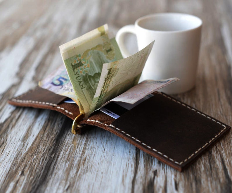 May include: A brown leather wallet with a metal money clip. The wallet is open and has Chinese Yuan bills inside. The wallet is sitting on a wooden table next to a white coffee mug.