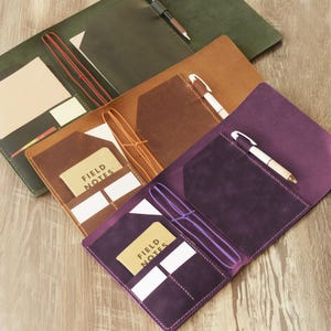 May include: Three leather notebook covers in dark green, tan, and purple. Each cover features a pocket, an elastic band for securing a notebook, and a pen holder containing a pen. A Field Notes brand notebook is visible in the tan and purple covers.
