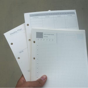 May include: A white weekly planner with a gridded page and a section for appointments. The planner has a tab that says "Weekly Plan" and a section for each day of the week. The planner has a hole punch on the left side.