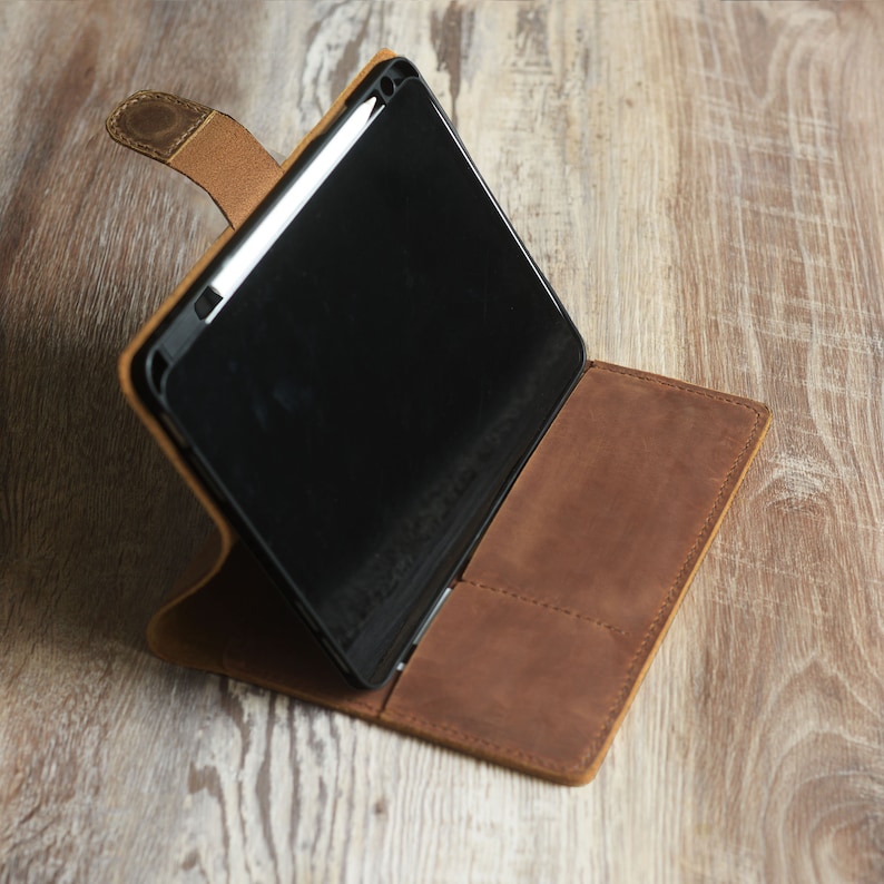 以下が含まれることがあります： 黒いタブレットと白いスタイラスが収納された茶色の革製ケース。ケースが開いており、タブレットが見えています。