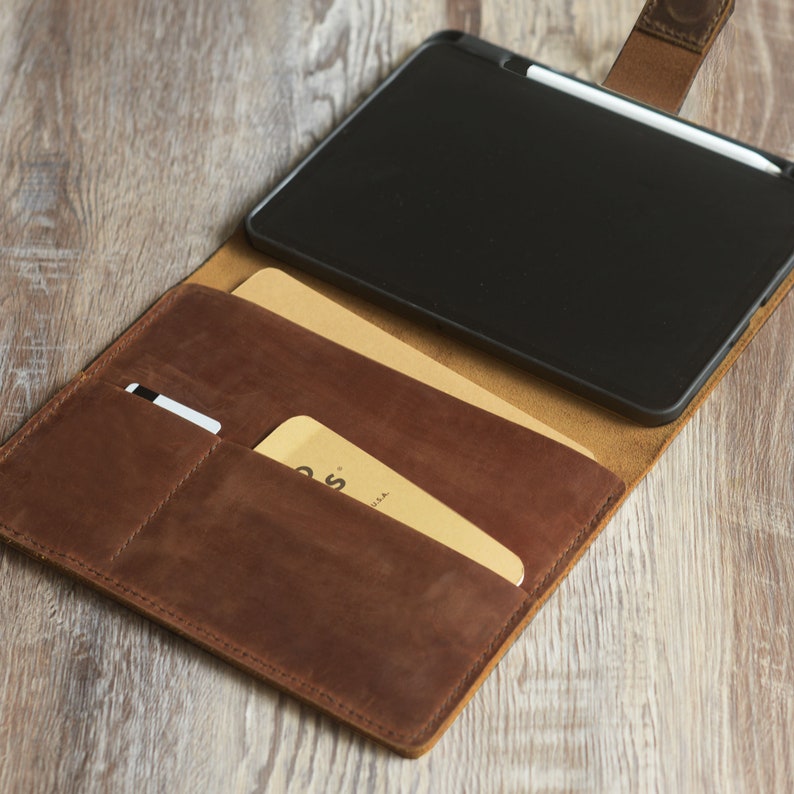 以下が含まれることがあります： 黒いタブレットとスタイラスが入った茶色の革製フォリオケース。ケースにはカードやその他のアイテムを入れるためのポケットが2つあります。