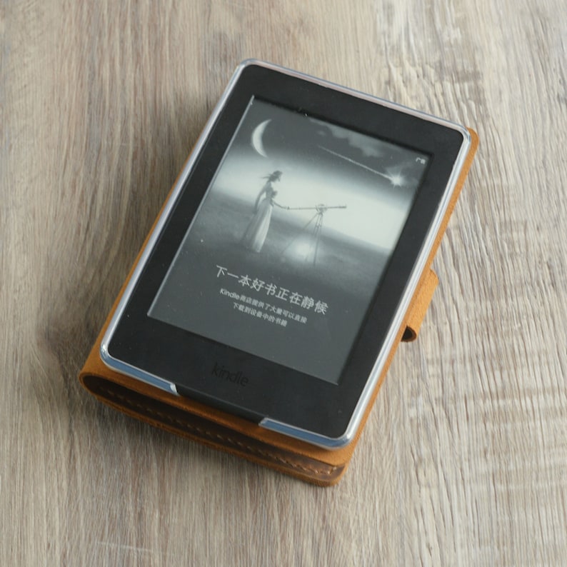 Peut inclure: Un &eacute;tui en cuir marron avec une bordure argent&eacute;e entoure une liseuse Kindle noire. L'&eacute;cran affiche une image en noir et blanc d'une femme debout dans un champ avec un t&eacute;lescope. Le texte &agrave; l'&eacute;cran indique "下一本好书正在静候 Kindle提供了大量可以下载到设备中的书籍".