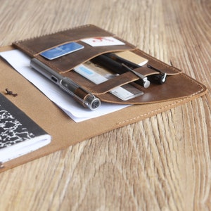 May include: A brown leather portfolio with multiple pockets and slots for holding pens, cards, and a notebook. The portfolio is open and the notebook is visible.