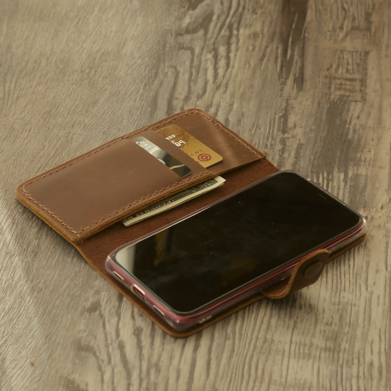 Peut inclure: Un &eacute;tui de t&eacute;l&eacute;phone en cuir marron avec un emplacement pour carte int&eacute;gr&eacute; et un rabat qui se ferme &agrave; l'aide d'un bouton. L'&eacute;tui est ouvert et un smartphone est visible &agrave; l'int&eacute;rieur. Il y a deux cartes de cr&eacute;dit et de l'argent liquide dans l'emplacement pour carte.