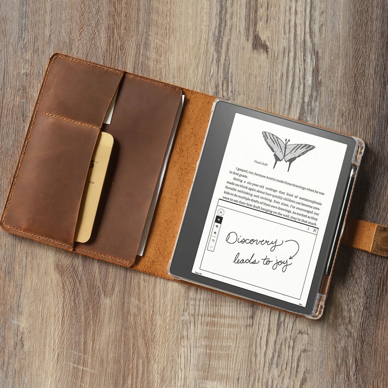 Op de afbeelding: Een bruine leren folio-hoes met een ingebouwde pocket voor een stylus en een aparte pocket voor een klein notitieboekje. De hoes is open en een zwarte e-reader bevindt zich erin. Het e-reader-scherm toont een vlinderillustratie en de tekst "Discovery? leads to joy".