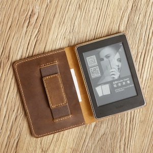 Peut inclure: Un &eacute;tui en cuir marron avec une poche pour une carte de cr&eacute;dit ou d'autres petits objets. L'&eacute;tui est ouvert pour r&eacute;v&eacute;ler une liseuse Kindle Oasis noire avec un &eacute;cran affichant une image promotionnelle pour l'appareil. Le texte sur l'&eacute;cran indique "Kindle Oasis, 现在, 敢读 | 敢夢".