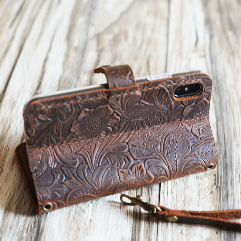 K&ouml;nnte beinhalten: Eine braune Leder Handyh&uuml;lle mit einem gepr&auml;gten Blumenmuster. Die H&uuml;lle hat eine Klappe, die mit einem Druckknopf geschlossen wird, und einen Riemen mit einem Metallverschluss.