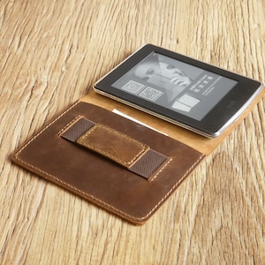 May include: A brown leather case with a flap that folds over to protect an e-reader. The case has a built-in elastic strap to hold the e-reader in place. The e-reader is visible with the screen displaying a promotional image for Kindle.