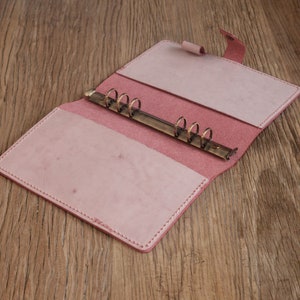Op de afbeelding: Een roze lederen gebonden dagboek met een messing ringband. Het dagboek is open om de lege pagina's binnenin te laten zien.