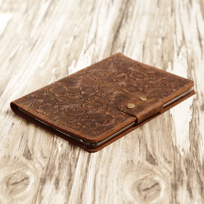 May include: A brown leather tablet case with a floral embossed design. The case has two brass snaps on the front.