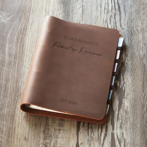 Puede incluir: Un libro de recetas de cuero marrón con un acabado mate suave. La cubierta está grabada con el texto CORA & JAKEB'S Family Recipes y EST 2026. El libro cuenta con múltiples separadores de pestañas de colores que sobresalen por el lado derecho.