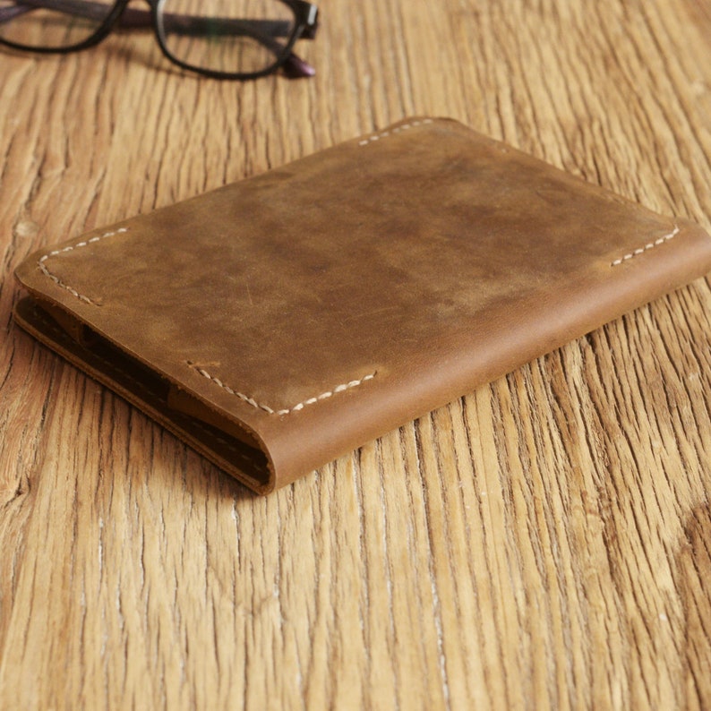 Puede incluir: Un cuaderno de cuero marr&oacute;n con costuras blancas. El cuaderno est&aacute; cerrado y sobre una superficie de madera.