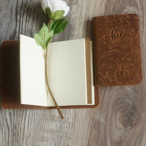 May include: Two brown leather journals with a floral embossed design. One journal has the text "her vows" and "EMMA 8.11.22" on the cover. The other journal is open and has blank pages.