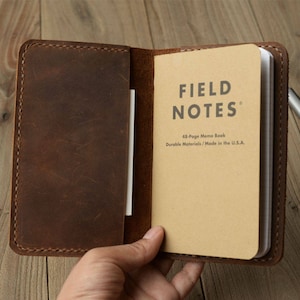 May include: A brown leather notebook cover with a Field Notes 48-page memo book inside. The book has the text "FIELD NOTES" and "48-Page Memo Book" and "Durable Materials / Made in the U.S.A."