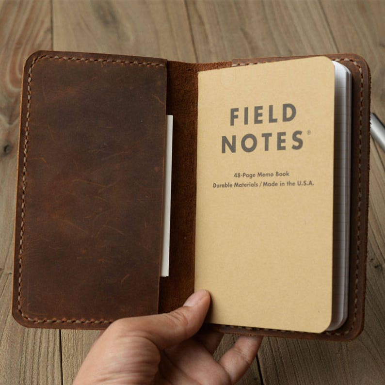 May include: A brown leather notebook cover with a Field Notes 48-page memo book inside. The book has the text "FIELD NOTES" and "48-Page Memo Book" and "Durable Materials / Made in the U.S.A."