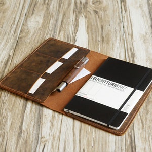May include: A brown leather notebook cover with a black notebook inside. The notebook has the text "LEUCHTTURM1917" and "DETAILS MAKE ALL THE DIFFERENCE." The cover has multiple slots for cards and a pen holder.